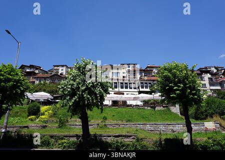 Smolyan, Bulgaria, Una pittoresca cittadina collinare con un mix di architettura tradizionale e moderna. La città è adagiata su una collina in pendenza, con un cielo azzurro. Gli edifici sono principalmente bianchi e beige con alcuni tetti di tegole rosse, e alcuni hanno balconi. In primo piano sono visibili un muro di contenimento in pietra e un'area erbosa con alberi e arbusti. Diversi ombrelli bianchi sono disposti su un'area patio di fronte ad un edificio con una facciata color crema o beige e persiane marrone scuro o nere. Sul lato sinistro dell'immagine è visibile anche una luce stradale. Occhio e immaginazione di un artista Foto Stock