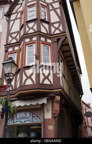 Wertheim, alla confluenza dei fiumi Main e Tauber nel Baden-Württemberg, Germania. Un edificio ad angolo con una facciata a metà legno, caratterizzato da una combinazione di colori marrone e crema/beige. L'edificio ha finestre multiple con cornici bianche e finiture arancioni/marroni, e un piccolo negozio con una tenda bianca si trova all'angolo. Un lampione si trova sul lato sinistro dell'immagine e una porzione di un altro edificio è visibile sulla destra. Lo stile architettonico dell'edificio ricorda il tradizionale mezzo legno di legno Foto Stock