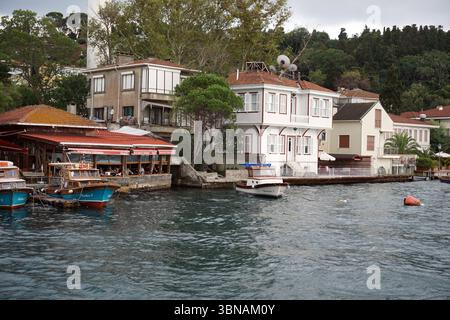 Istanbul, Turchia. La città più grande della Turchia e una delle poche città transcontinentali del mondo, che si estende sia in Europa che in Asia attraverso lo stretto del Bosforo, Una pittoresca scena sul lungomare con una fila di case ed edifici lungo la costa. Le case sono dipinte in una varietà di colori, tra cui bianco, rosso e beige, e presentano tetti di tegole rosse. Alcune delle case hanno balconi e piatti satellitari. Le case sono situate su una collina, con lussureggianti alberi verdi e una collina coperta di vegetazione sullo sfondo. L'acqua è di colore blu-verde scuro e lungo le quali sono attraccate diverse barche Foto Stock
