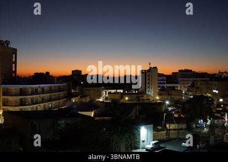 Larnaca, Cipro, l'immagine cattura un paesaggio urbano al crepuscolo, con un cielo arancione e blu vibrante che si trasforma in un blu profondo. L'area e' illuminata dal caldo bagliore di lampioni e luci dell'edificio, creando un contrasto tra le sagome scure degli edifici e le luci luminose. Gli edifici sono per lo più bassi, con un mix di stili architettonici, e sono immersi nella luce soffusa del sole che tramonta. Il paesaggio urbano è visto da un alto punto panoramico, offrendo una vista panoramica del paesaggio urbano. L'immagine viene acquisita da un angolo alto, fornendo una vista completa del layout e. Foto Stock