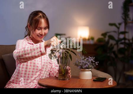 Giovane donna asiatica adulta in pigiama rosa che innaffia piante al coperto come pratica di vita di longevità per connettersi con la natura migliorando il benessere per l lento Foto Stock