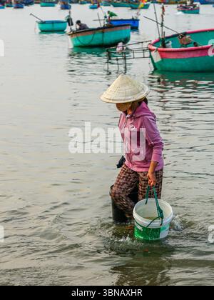 MUI ne, Vietnam - 22 marzo 2025: Una pescatrice locale a Mui ne, torna a riva con secchi con pescato fresco, circondato da tonde di pescherecci Foto Stock