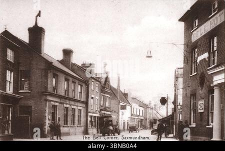 Una tranquilla scena di strada a Saxmundham, East Suffolk, centrata sul Bell Hotel, un prominente edificio in mattoni con un cartello sospeso. Alcuni pedoni si soffermano vicino all'ingresso e i primi veicoli a motore, probabilmente dalla fine degli anni '1910 all'inizio degli anni '1920, sono parcheggiati lungo il marciapiede. La strada è costeggiata da camini e negozi, riflettendo la modesta prosperità di una città mercato negli anni tra le due guerre. Da una cartolina originale, non attribuita. Foto Stock