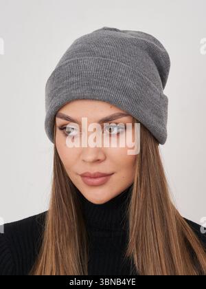 La donna indossa un berretto in maglia grigia con capelli dritti marroni visibili, vestito con dolcevita nero, che guarda fuori dalla fotocamera. Toni terrosi, espressione concentrata Foto Stock