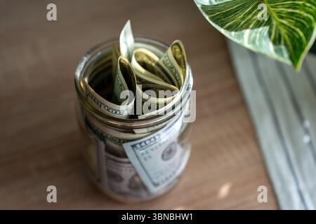 Vaso di vetro pieno di banconote in dollari USA arrotolate su un tavolo di legno con luce naturale. Foto Stock