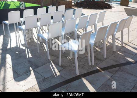 File di moderne sedie in plastica bianca sono disposte all'aperto su un patio in pietra illuminato dal sole, che proietta forti ombre. Ideale per eventi e riunioni Foto Stock