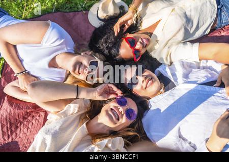 Quattro diverse giovani donne sdraiate insieme in un parco su una coperta con occhiali da sole e sorridenti alla macchina fotografica Foto Stock