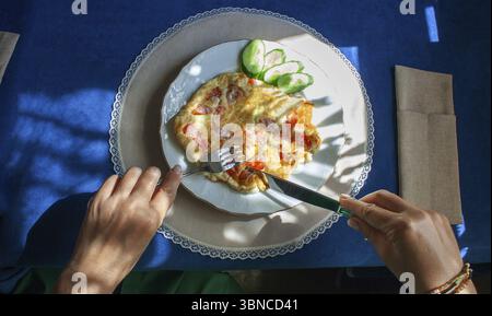 Le mani femminili tagliano uova strapazzate con pomodori, su cui cade l'ombra di una tenda con le nappine, colazione in una giornata di sole Foto Stock