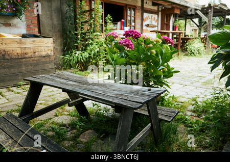 Il tavolo da picnic in legno si trova di fronte a un giardino fiorito. Il tavolo è vuoto e i fiori sono rosa Foto Stock