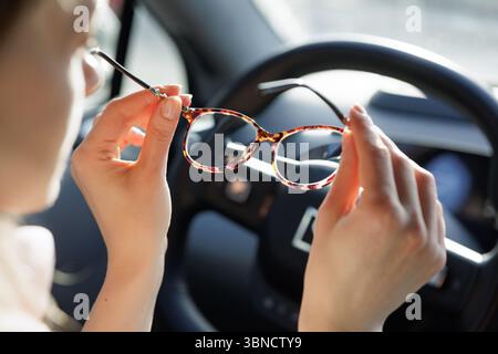 Primo piano di una donna che tiene gli occhiali all'interno dell'auto, luce naturale che mette in risalto la chiarezza visiva e guida sicura. Foto Stock