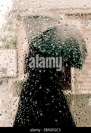 Una persona cammina sotto la pioggia, tenendo in mano un ombrello. L'immagine viene scattata da dietro una finestra coperta di gocce di pioggia. Foto Stock
