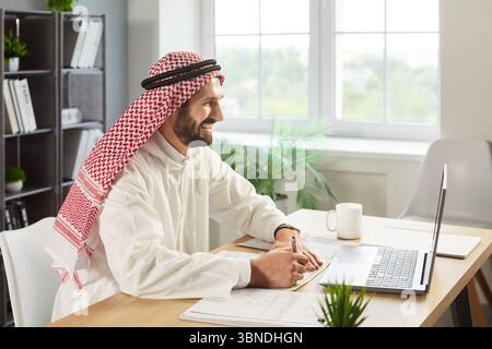 Uomo arabo sorridente in kandura bianca seduto alla scrivania, utilizzando il computer portatile e prendendo appunti durante la videochiamata. Foto Stock