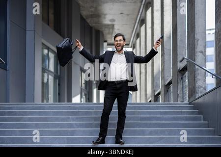 Un uomo d'affari con una tuta festeggia con le braccia alzate, tenendo una valigetta e uno smartphone sulle scale all'aperto, sembrando entusiasta. Foto Stock