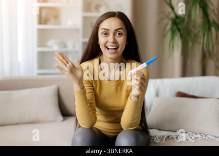 Donna felicissima che tiene il test di gravidanza con le mani, sorride e solleva la mano Foto Stock
