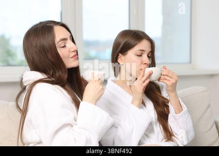 Belle donne in accappatoi con tazze di bevanda sana dopo le procedure spa sul divano al chiuso Foto Stock