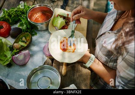 Una persona prepara tacos utilizzando ingredienti freschi, tra cui salsa, verdure e tortillas, su un tavolo rustico all'aperto, evocando freschi e avventurosi Foto Stock