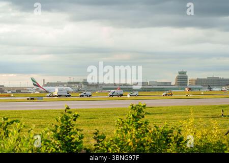 Una vista panoramica della pista dell'aeroporto di Manchester con veicoli operativi in preparazione alla manutenzione e aerei visibili in una serata nuvolosa. Foto Stock
