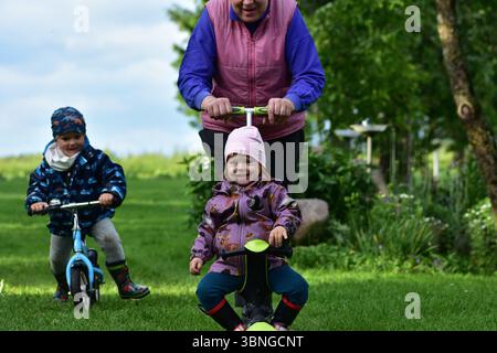 Una ragazza allegra con un cappello rosa viene spinta da sua nonna su uno scooter, mentre un bambino sorridente guida una bicicletta sullo sfondo. Divertimento per tutta la famiglia a gre Foto Stock