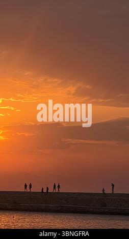 Sagome di persone che camminano e si alzano su un molo durante uno splendido tramonto arancione. Il cielo è pieno di sfumature vibranti, creando un ambiente sereno e pictu Foto Stock