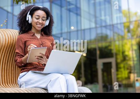 Una donna sorridente che indossa cuffie e occhiali si siede all'esterno, prendendo appunti su un notebook e lavorando su un notebook. Foto Stock
