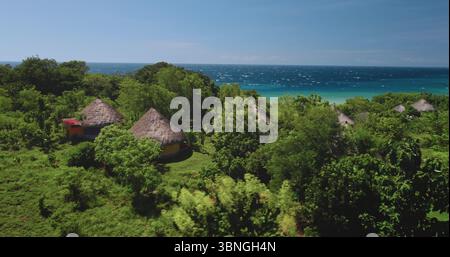 Tradizionali case indonesiane con tetti di paglia accoccolate tra una vibrante vegetazione verde sull'Isola di Sumba, che si affaccia sul turchese Oceano Indiano sotto un cielo azzurro Foto Stock