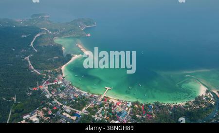 Vista aerea che cattura una strada costiera che si snoda attraverso una vegetazione lussureggiante, che conduce a una vivace città accanto a una pittoresca baia con acque cristalline e turchesi sull'isola di Koh Kood Foto Stock