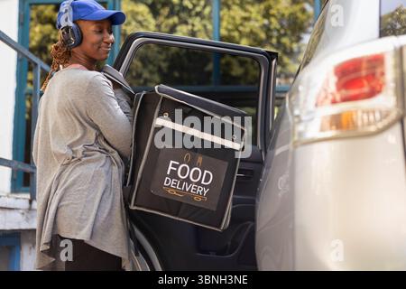 Il lavoratore da asporto esce dall'auto con lo zaino per consegnare il cibo, consegnando il pasto da un ristorante affollato. Servizio rapido per ordinare rapidamente i cibi da asporto all'ingresso. Foto Stock