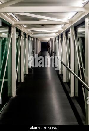 Un corridoio vuoto e ben illuminato con pavimento scuro e travi di supporto bianche. Il corridoio ha pareti di vetro su entrambi i lati e un corrimano sul lato destro. Foto Stock