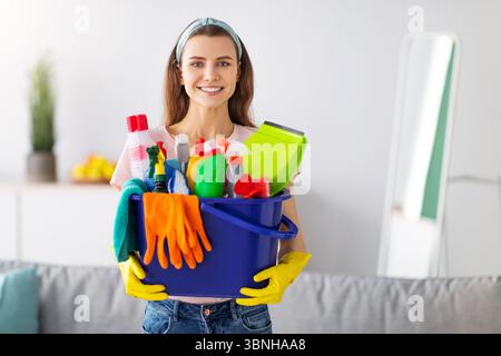 Gioiosa giovane cameriera che tiene il secchio pieno di rifornimenti di pulizia a. soggiorno Foto Stock