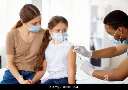 Madre e figlia in maschere mediche che si vaccina contro covid-19, medico che dà iniezione di vaccino del coronavirus Foto Stock