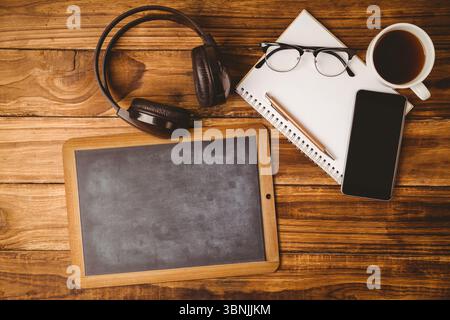 Il tablet vuoto in stile lavagna si appoggia su una scrivania in legno e mostra le cuffie, lo smartphone e il caffè Foto Stock