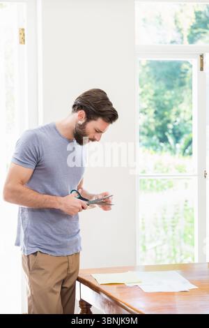 Forbici che tagliano la carta su un tavolo di legno con fogli sparsi accanto alla finestra illuminata dal sole che mostra il giardino verde Foto Stock
