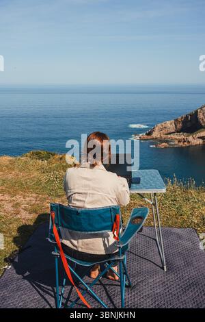 Una donna ama lavorare a distanza sedendosi su una sedia vicino all'oceano, circondato da viste mozzafiato. Una configurazione ideale per i nomadi digitali che viaggiano in videocamera Foto Stock