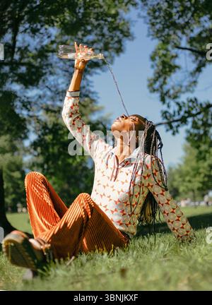 Una donna afroamericana con i capelli intrecciati ama l'acqua in natura, seduto sull'erba in un parco illuminato dal sole. Versa l'acqua da una bottiglia con grazia, apprecia Foto Stock