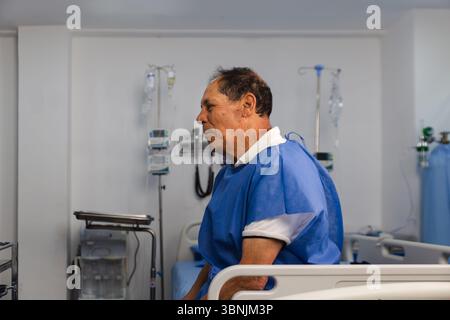 Un uomo anziano in una stanza d'ospedale che indossa un abito blu. Si siede su un letto d'ospedale, circondato da attrezzature mediche, mostrando un expres calmo e sereno Foto Stock