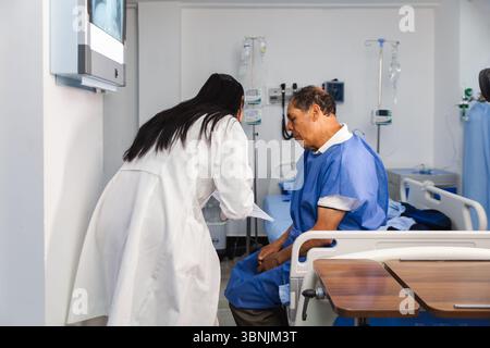 Un medico donna in un cappotto bianco si consulta con un paziente anziano di sesso maschile seduto su un letto d'ospedale, discutendo informazioni mediche in un luminoso e ben attrezzato Foto Stock