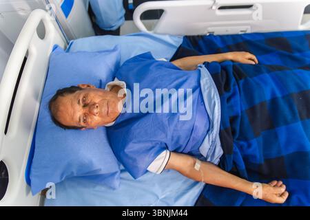 An elderly man lies in a hospital bed, comfortably resting under a blue checkered blanket. He appears calm and relaxed, suggesting a peaceful recovery Foto Stock