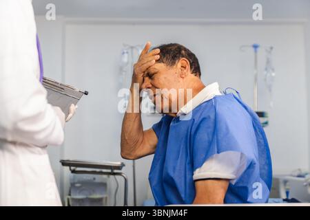 Un paziente anziano in un abito blu siede in una stanza d'ospedale, tenendo la testa in dolore mentre si consulta con un medico donna, che tiene un portadocumenti durino Foto Stock