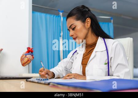 Un medico dedicato in un cappotto bianco scrive appunti a una scrivania in uno studio medico. Un modello di cuore è visibile, significa impegno sanitario e professionale Foto Stock