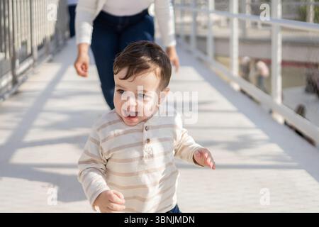Un bambino allegro esplora felicemente il parco mentre sua madre lo segue da vicino, godendosi una giornata di sole insieme. L'atmosfera luminosa riflette un gioioso e bo Foto Stock