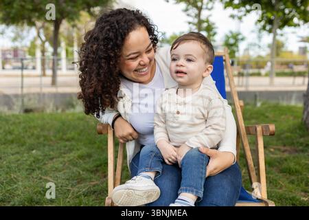 Una madre gioiosa e il suo bambino si godono una giornata di sole nel parco, condividendo risate e creando ricordi preziosi seduti su una sedia di legno circondata da lu Foto Stock