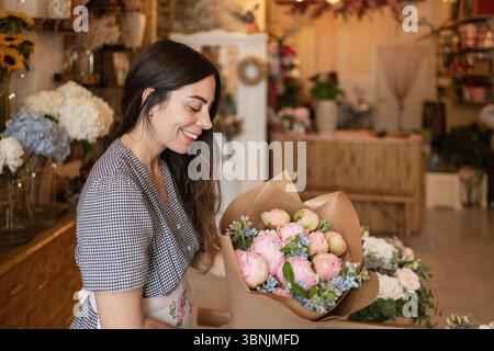 Una donna organizza un bouquet di fiori rosa e blu in un affascinante negozio di fiori. Sorride, indossa una camicetta a scacchi e un grembiule floreale, circondato da co Foto Stock