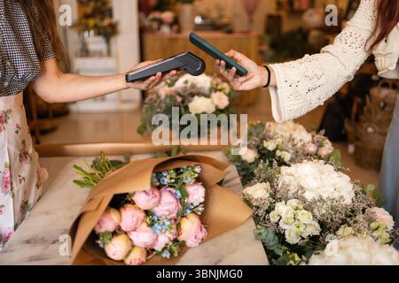 Le donne in un'azienda fiorentina utilizzano pagamenti senza contatto su dispositivi mobili circondati da un assortimento di mazzi di fiori colorati, che esemplificano i moderni ret Foto Stock