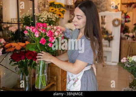 Una donna organizza rose rosa vivaci in un vaso di vetro in un'affollata fioreria. Circondata da fiori colorati, crea con cura un bellissimo disp floreale Foto Stock