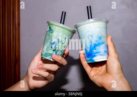 Due mani per frullati verdi e blu vivaci in tazze da asporto trasparenti con cannucce nere. Queste bevande rinfrescanti incarnano uno stile di vita sano e sono perf Foto Stock