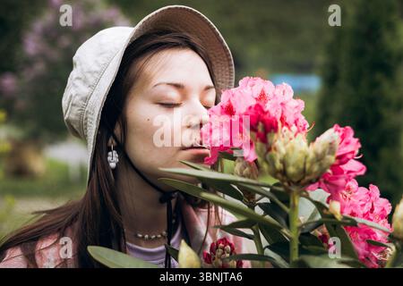 Una donna con un cappello da sole gode del profumo di rododendri rosa vibranti in un tranquillo giardino, che illustra un momento tranquillo di connessione con na Foto Stock