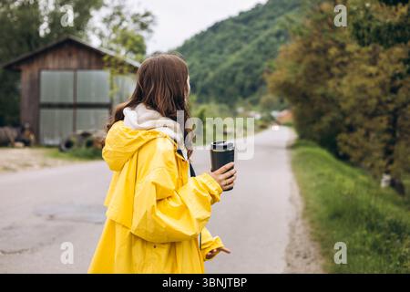 Una giovane donna si erge tra le tranquille montagne, indossando un impermeabile giallo brillante, godendosi l'aria fresca e viste mozzafiato, mentre tiene in mano una tazza da viaggio Foto Stock
