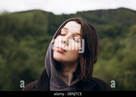 Una donna serena sta con gli occhi chiusi, abbracciando la tranquillità della natura. Indossa una felpa con cappuccio, circondata da verde lussureggiante e gentile Foto Stock