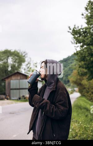 Una giovane donna con un abbigliamento informale si gode una bevanda calda da una tazza thermos mentre si trova all'aperto. La scena intima è ambientata sullo sfondo della lussureggiante greene Foto Stock