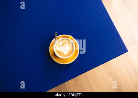 Vista dall'alto verso il basso di una tazza di caffè con latte art a forma di cigno. La tazza giallo brillante contrasta con il ricco tavolo blu, creando un vivace e invitante caffè Foto Stock